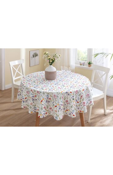 Runder Esstisch mit weißer Tischdecke, buntes Blumenmuster, überhängend, zwei weiße Stühle mit Kreuzlehne, weiße Vase mit Blumen in der Tischmitte, helles Zimmer mit Fenster und Wandbild