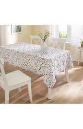 Rechteckige Tischdecke mit buntem Blumenmuster in Rot, Blau, Gelb und Grün, auf weißem Untergrund, liegt auf Holztisch, umgeben von weißen Holzstühlen, in hellen Raum mit Fensterlicht
