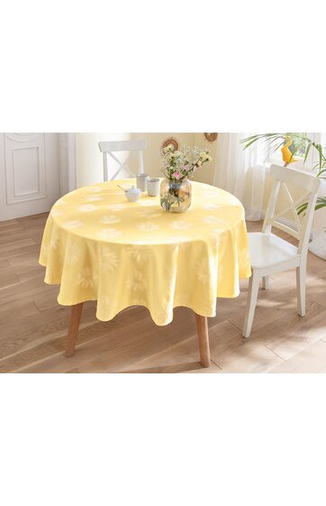 Runder Esstisch mit gelbem Tischdecke, floral geprägtes Muster, helle Holzbeine, darauf Glasvase mit gemischten Blumen, zwei weiße Stühle, heller Raum mit Fenster und grünem Pflanzenblatt