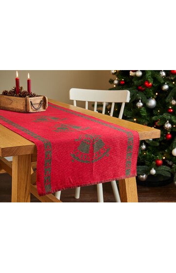 Roter Tischläufer mit grünem Weihnachtsglockenmotiv und grünem Randmuster, liegt auf einem hellen Holztisch, im Hintergrund weiße Holzstühle und Dekoration mit Weihnachtsbaum und roten Kugeln