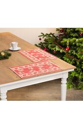 Zwei rechteckige Tischsets in rot und beige mit Weihnachtsbaum- und Sternmotiven, flach liegend auf Holztisch mit weißem Untergestell, neben geschmücktem Tannenbaum, weiße Kaffeetasse mit Untertasse auf Tisch