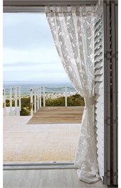 Weißer Gardinenvorhang mit floralem Muster, halb geöffnet und an der rechten Seite zusammengebunden, Blick auf Terrasse mit Holzdeck, weißem Geländer und weiter Landschaftshorizont im Hintergrund