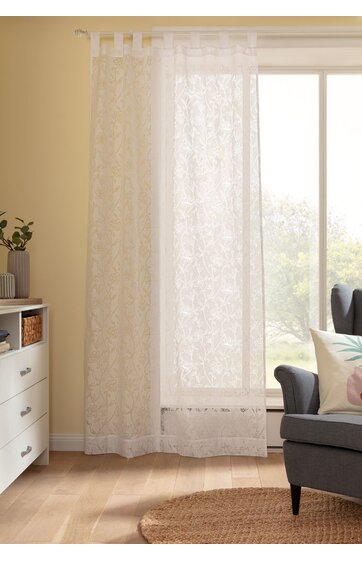 Transparenter, weißer Gardinenvorhang mit floralem Muster, lang, an weißen Ösen befestigt, hängend vor großem Fenster, im Wohnzimmer neben grauem Sessel und weißer Kommode auf hellem Holzboden
