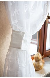 Weiße Gardine mit Muster aus Kreuzlinien, zusammengebunden mit weißem Stoffband mit Knopf, vor Fenster mit braunem Holzrahmen, Fensterbank mit Dekoration
