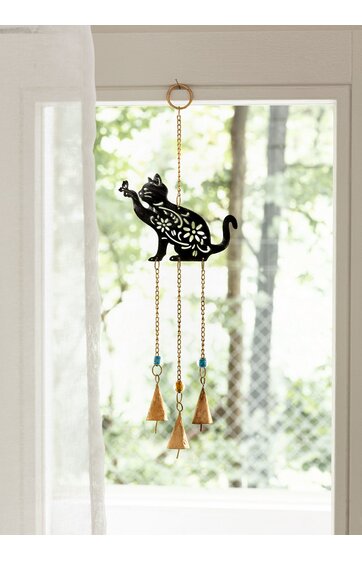 Windspiel mit schwarzer Katzenfigur, filigranes Blumenmuster, aufgehängt an goldener Kette, drei hängende goldfarbene Glocken und blaue Zierringe, vor hellem Fenster mit grünem Baumhintergrund
