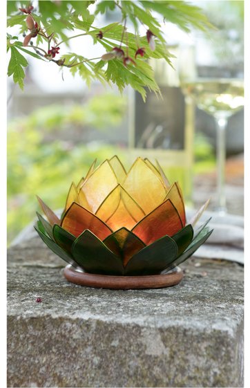 Teelichthalter in Form einer aufblühenden Blume, grüne und gelbe Glasblätter, mehrschichtig angeordnet, auf rundem braunem Untersetzer, steht auf Steinfläche im Freien, unscharfer Hintergrund mit Pflanzen und Glasgefäß