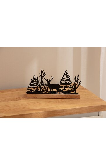 Schwarze Holzdekoration mit Rentier, zwei Tannenbäumen und zwei kahlen Büschen auf braunem rechteckigem Sockel, platzierte auf hellem Holztisch vor beiger Wand