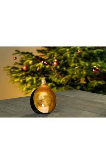 Tischkerze in goldener, runder Laterne mit Glasfenster, Kerzenlicht sichtbar, auf grauem Tisch, unscharfer grüner Tannenbaum mit roten Kugeln im Hintergrund