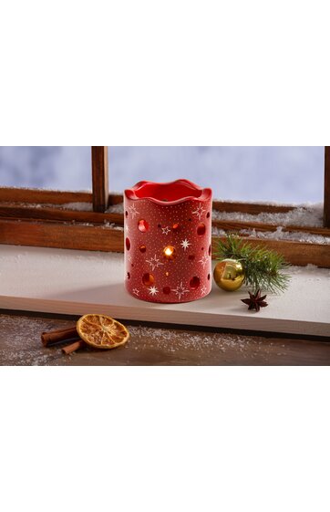 Rotes Windlicht mit sternenförmigen und runden Ausstanzungen, gewellte obere Kante, kerzenlichtscheinend, auf weißem Untergrund neben getrockneter Orangenscheibe, Zimtstangen, grüner Tannenzweig und goldener Weihnachtskugel vor Holzfenster mit Schnee