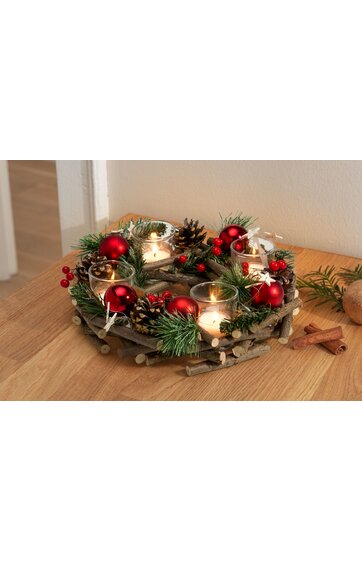 Adventskranz aus dunklem Holz mit grünen Tannenzweigen, roten Weihnachtskugeln, Tannenzapfen, roten Beeren, vier kleinen weißen Kerzen in Glashaltern, auf hellem Holztisch stehend