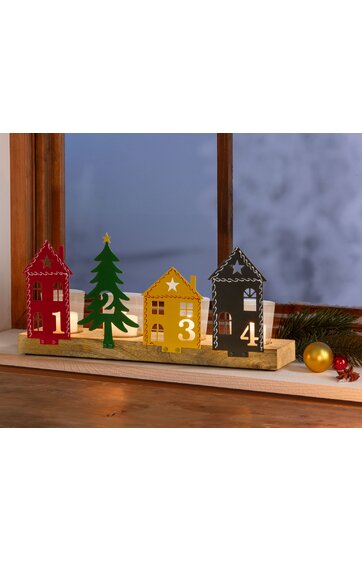 Adventskalender aus Holz mit vier Reihen, bestehend aus roten, grünen, gelben und schwarzen Häuschen mit den Zahlen 1 bis 4, grüne Tannenbaumfigur mit Stern, auf hellem Holzsockel stehend, Dekoration auf Holztisch vor Fenster