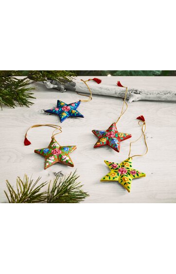 Vier sternförmige Weihnachtsanhänger in blau, rot, grün und gelb mit bunten Blumenmustern, jeweils mit goldfarbener Schlaufe und rotem Quast zum Aufhängen, flach liegend auf hellem Holzuntergrund, mit Tannenzweigen im Bild