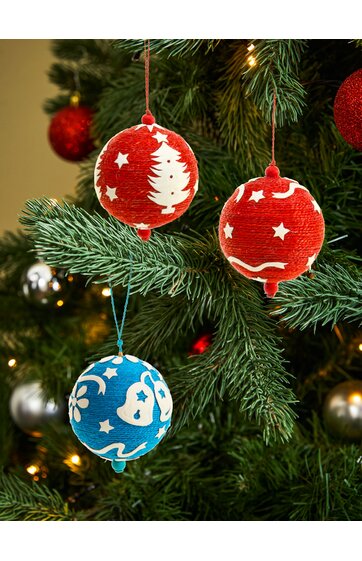 Drei Christbaumkugeln zum Aufhängen, zwei rot mit weißen Sternen und Tannenzapfenmotiven, eine blau mit weißen Blumenmustern, an grünen Tannenzweigen hängend