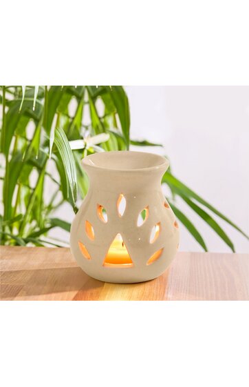 Weiße Keramik-Duftlampe mit Blätter- und Tropfenförmigen Aussparungen, runde Form, mittlere Öffnung am oberen Rand, beleuchtet, steht auf hellem Holztisch vor grünen Pflanzen