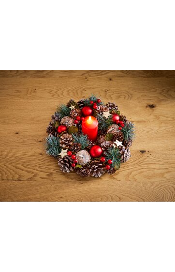 Adventskranz aus Tannenzweigen, roten Beeren und Zapfen, mit roten Kugeln und kleinen Sternen, rotes zylindrisches Bratapfelkerze in der Mitte, auf hellem Holzuntergrund