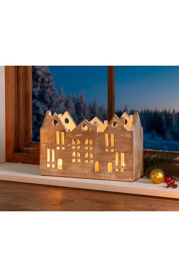 Holzdekoration in Form eines Hauses mit mehreren Gauben und beleuchteten rechteckigen Fenstern, braun, mit warmem Licht von innen, steht auf Fensterbank vor Schneelandschaft bei Dämmerung