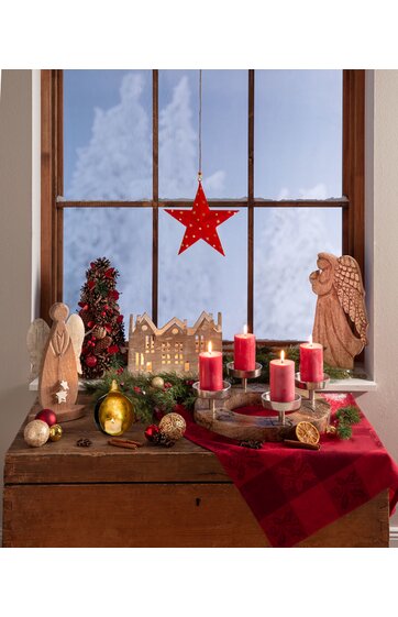 Weihnachtliche Dekoration auf dunklem Holztisch vor Fenster mit Himmel, roter Stern aus Papier hängend, zwei hellbraune Holzengel, kleine Tannenbaumfigur mit roten Kugeln, beleuchtetes Holzhaus, rote Adventskerzen auf Holzkranz, grüne Tannenzweige, r