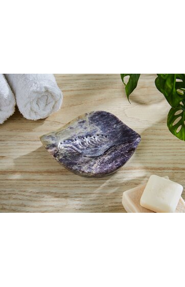 Seifenablage in organischer, wellenförmiger Form, mehrfarbig in violett und beige mit eingraviertem Blattmotiv in der Mitte, steht auf hellbraunem Holzuntergrund, daneben weiße Handtücher und zwei weiße Seifenstücke