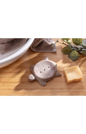 Fischförmiger Seifenspenderhalter in graubraun, flach, mit drei Löchern auf der Oberseite, rundem Kopf und Flossen, liegend auf hellem Holzbrett neben einem quadratischen gelben Schwamm und Handtüchern