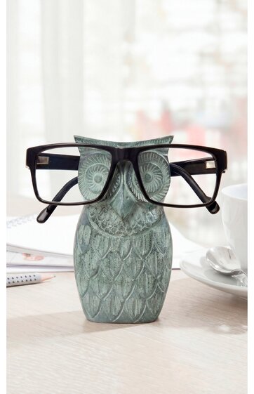Brillenhalter in Eulenform, graugrün mit geprägtem Federdesign, große schwarze rechteckige Brille auf dem Eulenkopf platziert, steht auf weißer Tischoberfläche neben weißer Tasse und Schreibblock