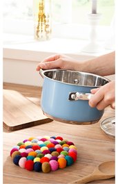 Blauer Kochtopf mit silbernem Innenraum und Edelstahlgriff, gehalten von Händen über einem Holztisch, bunte runde Filzunterlage aus kleinen Kugeln, Holzkelle und Holzbrett auf dem Tisch