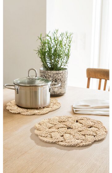 Runder beiger Topfuntersetzer aus geflochtenem Material auf hellem Holztisch, Edelstahl-Topf mit Glasdeckel darauf, weiterer Untersetzer im Vordergrund, grünes Kräuterpflanze in transparentem Blumentopf, Holzstuhl und weiße Gardine im Hintergrund