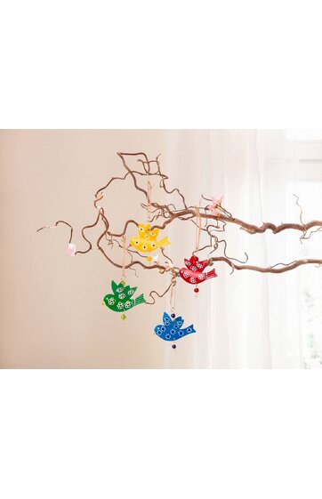 Vieranhänger in Vogelform in Gelb, Rot, Blau und Grün mit weißen Blumenmustern, hängend an verdrehtem braunem Ast vor hellem Fenstervorhang