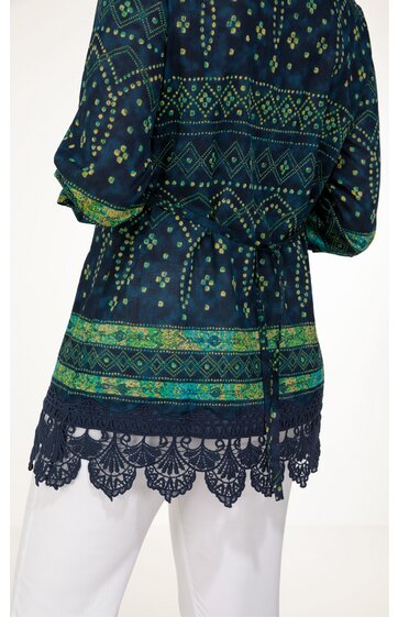 Dunkelblaue Tunika mit grünem geometrischem Muster, Raglanärmel, Taillenzug mit Kordel, Saum mit schwarzer Spitzenbordüre, getragen von Person in weißer Hose, locker geschnitten