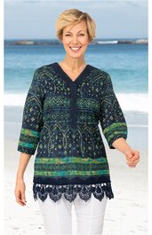 Dunkelblau-grüner Pullover mit mehrfarbigem Ethno-Muster, V-Ausschnitt mit Knopfleiste, 3/4-Ärmel, gehäkelter Spitzenabschluss, getragen von lächelnder stehender Frau vor Strandkulisse