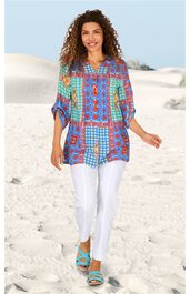 Locker sitzende Tunika in Blau, Türkis und Rot mit geometrischem Muster, V-Ausschnitt, dreiviertellange Ärmel, gerade Saumlinie, getragen von lächelnder Frau stehend im Sand, lange weiße Hose, türkisfarbene Sandalen