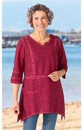 Damenpullover in dunklem Kirschrot, einfarbig, V-Ausschnitt mit Schlitz, verstellbare Taille mit Bindeband, 3/4-lange Ärmel mit feinen Strickdetails, asymmetrischer Saum, getragen von stehender lächelnder Frau am Strand
