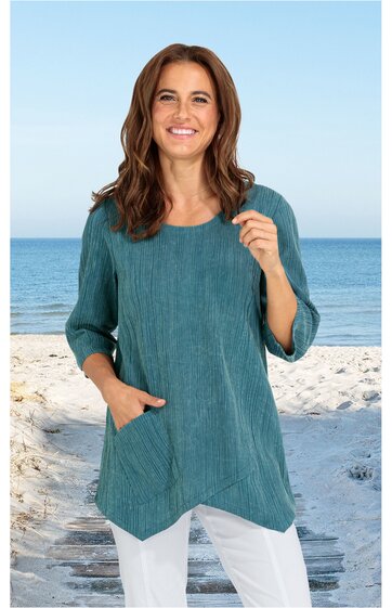 Locker sitzende Tunika in petrol mit feinem Streifenmuster, rundem Ausschnitt, drei Viertel Ärmeln, asymmetrischem Saum mit spitzen Enden, auf Hüfthöhe, getragen von lächelnder stehender Frau am Strand, zwei Taschen vorne