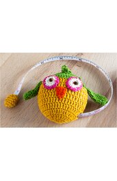 Gehäkeltes Spielzeug in Form einer Eule, gelb mit grünen Flügeln und Stacheln, rosa weiße große Augen, orangefarbener Schnabel, daneben liegendes Maßband mit gelbem Aufroller