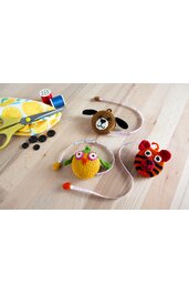 Drei handgehäkelte Maßbänder in Form eines Bärenkopfs braun-schwarz, einer Eule gelb-grün-pink und eines Tigers rot-schwarz, liegend auf hellem Holztisch mit gelber Schere, Nadelfaden und schwarzen Knöpfen im Hintergrund