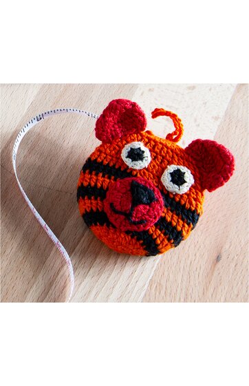 Kreisförmiges Maßband aus gehäkeltem orange-schwarzem Stoff, tigerähnliches Design mit roten Ohren, großen weißen Augen, roter Schnauze mit schwarzer Nase, auf hellem Holztisch liegend