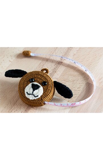 Rundes, braunes Maßband mit gehäkeltem Bezug in Form eines Hundekopfes, schwarzer Nase, schwarzem Mund, schwarzen Augen, zwei schwarzen Ohren, ausgezogenes weißes Band mit Zentimeter-Einteilung auf hellbraunem Holzuntergrund