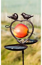 Metallfutterspender für Vögel in dunkelbraun, herzförmiger Rahmen, zwei stilisierte Vogelfiguren oben, mittig ein roter Apfel aufgespießt, darunter zwei runde Futterplatten mit Vogelsaat, freistehend im Garten