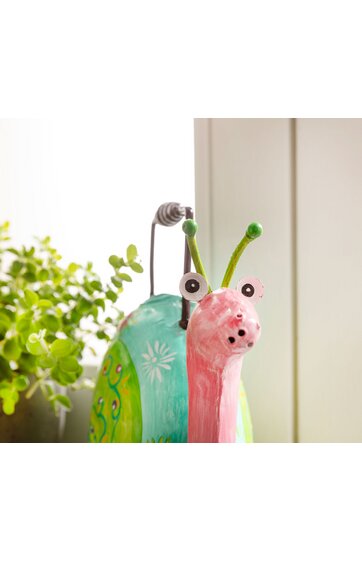 Bunte Schneckenfigur für Dekoration, rosa Kopf mit weiß-braunen Augen, grüne Fühler, türkis und grün gemusterter Körper mit weißen Blumenmotiven, steht auf Fensterbank neben grünem Pflanzenblatt