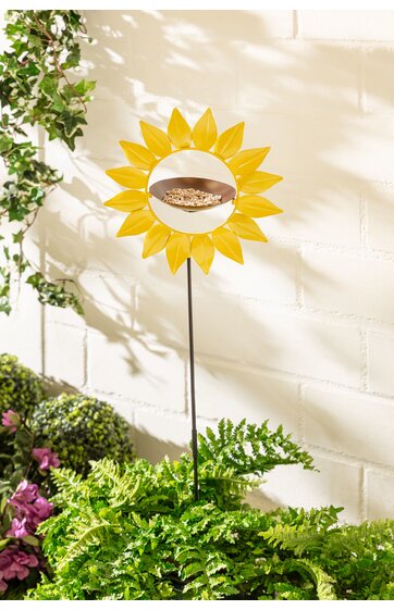 Gartendekoration Sonnenblume, gelb, rundes Design mit Öffnung in der Mitte, auf schlankem schwarzen Stab stehend, umgeben von grünen Pflanzen, vor hellen Mauersteinen