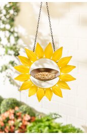 Gelbes Sonnenblumen-förmiges Vogelhaus aus Metall, rundes Futterschälchen in der Mitte, aufgehängt an Kette, sichtbare Körner im Futterschälchen, Hintergrund mit unscharfen grünen und weißen Elementen