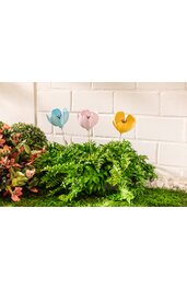 Drei bunte Tulpenstecker in Hellblau, Rosa und Gelb mit schwarzen Details, stehend in grünem Farnkraut, umgeben von weiteren grünen Pflanzen, vor weißer Klinkerwand