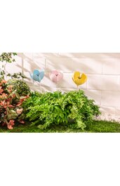 Drei bunte Blumenstecker in hellblau, rosa und gelb mit schwarzer Blütenzeichnung, steckend in grünem Farn, vor weißer Mauer, umgeben von weiteren grünen Pflanzen und Gras