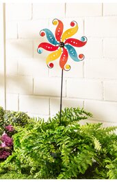 Gartendeko Windrad in Rot, Gelb, Blau mit schwarzen Punkten, sternförmiges Design mit gebogenen Blättern, montiert auf schwarzem Metallstab, platziert in grünem Farn vor weißer Wand