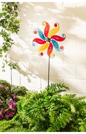 Gartenstecker mit mehrfarbigem Windrad in Rot, Gelb und Blau, drehbare Elemente, auf langem, dünnem schwarzem Stab stehend, umgeben von grünen Farnen und lila Blüten, vor heller Mauer