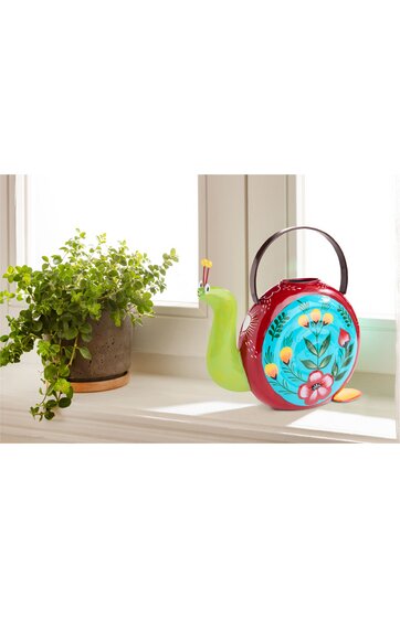 Gießkanne in Schneckenform, rot mit blauem Blumendekor, gelbe und orange Blütenmuster, grüner Schneckenkörper, runder Griff, auf Fensterbank neben grünem Blattpflanze in braunem Topf