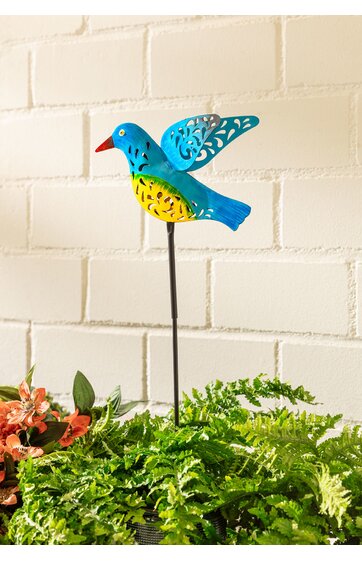 blauer Vogel auf Metallstab, gelber Bauch, rote Schnabelspitze, Flügel mit floralen Ausschnitten, steckbar, umgeben von grünen und orangefarbenen Pflanzen