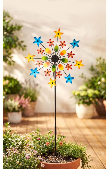 Gartendekoration mit Windspiel in Form eines Blumenrades, Ständer schwarz, Blütenblätter gelb, rot und blau, mittig schwarze Kreise, montiert in einem Pflanztopf mit grünen Pflanzen, Hintergrund unscharfe Topfpflanzen