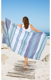 Großes rechteckiges Strandtuch in Blau-, Hellblau- und Weißtönen, gestreift, mit Fransen an den Enden, gehalten von einer stehenden Person am Strand, Rückansicht, Person trägt weißes Top und Gehen auf Holzplanken