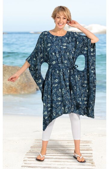 Damenkaftan in dunklem Blau mit floralem Muster in Weiß und Beige, lockere lange Fledermausärmel, Gürtel in Taillenhöhe, getragen von stehender Frau am Strand mit weißen Leggings und Flip-Flops