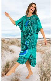 Kleid in türkis mit abstraktem dunkelblauem Muster, Rundhalsausschnitt, halblange lockere Ärmel, knielang, Figur umspielend, getragen von lachender Frau barfuß, stehend auf Sand mit Strandgras